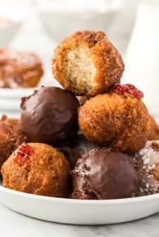 A plate of assorted donut holes, including chocolate-covered, powdered sugar, and jelly-filled varieties. The top cinnamon sugar donut hole has a bite taken out, revealing its fluffy interior.