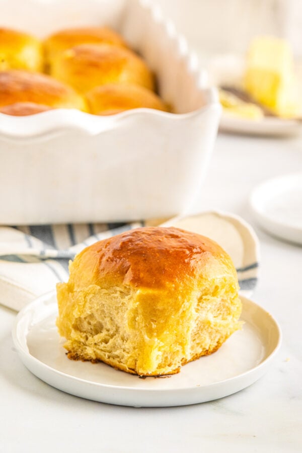 Soft & buttery Hawaiian Rolls are golden in color with a sweet hint of pineapple flavor. This homemade yeast roll recipe is a crowd favorite! A soft, tender and sweet Hawaiian roll on a small white plate, with the pan of rolls and a dish of butter in the background of the shot.