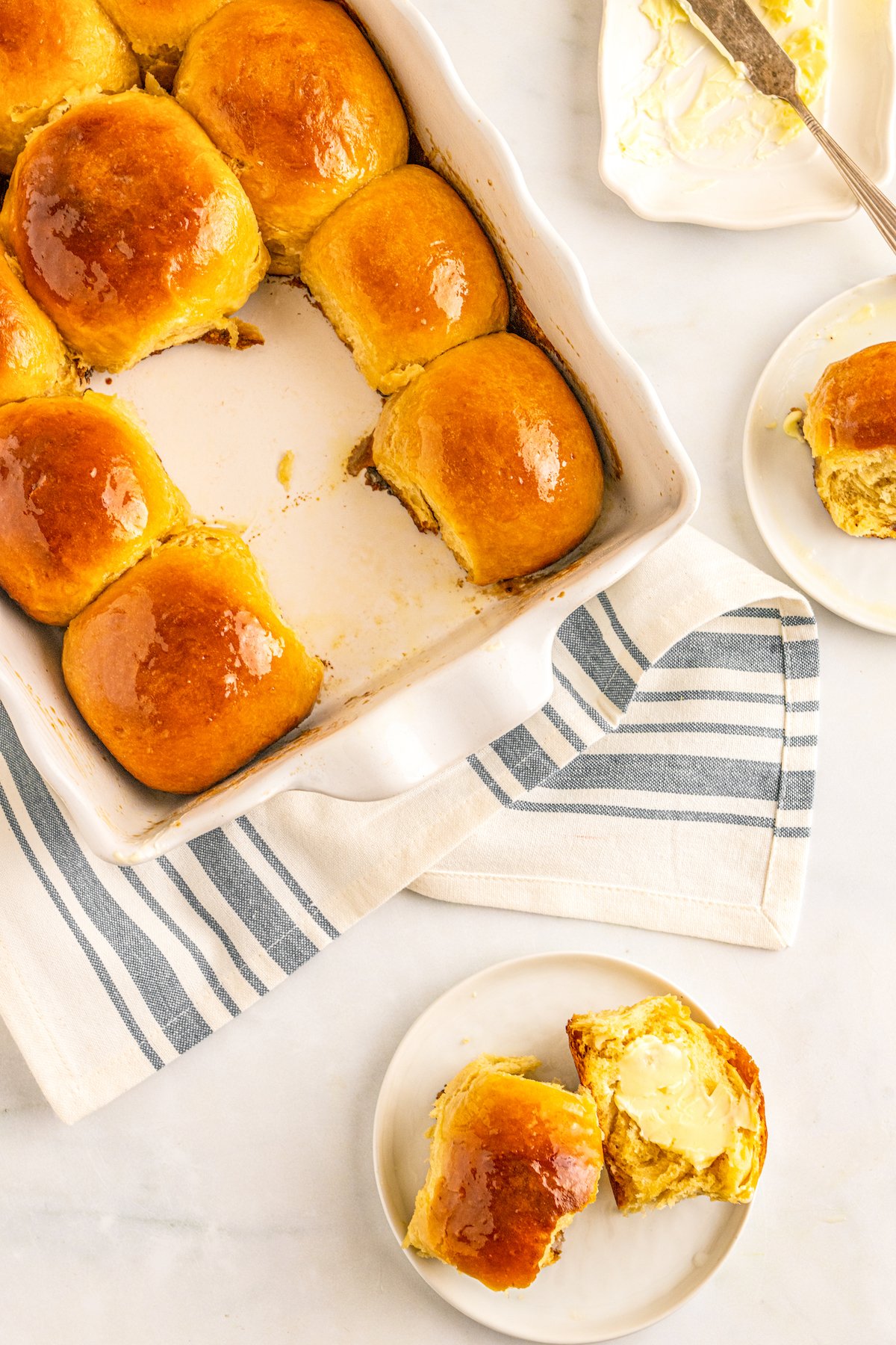 Soft & buttery Hawaiian Rolls are golden in color with a sweet hint of pineapple flavor. This homemade yeast roll recipe is a crowd favorite! Overhead shot of a table with a white baking dish of hawaiian bread rolls on a cloth napkin. Three of the rolls have been removed from the pan to small plates.