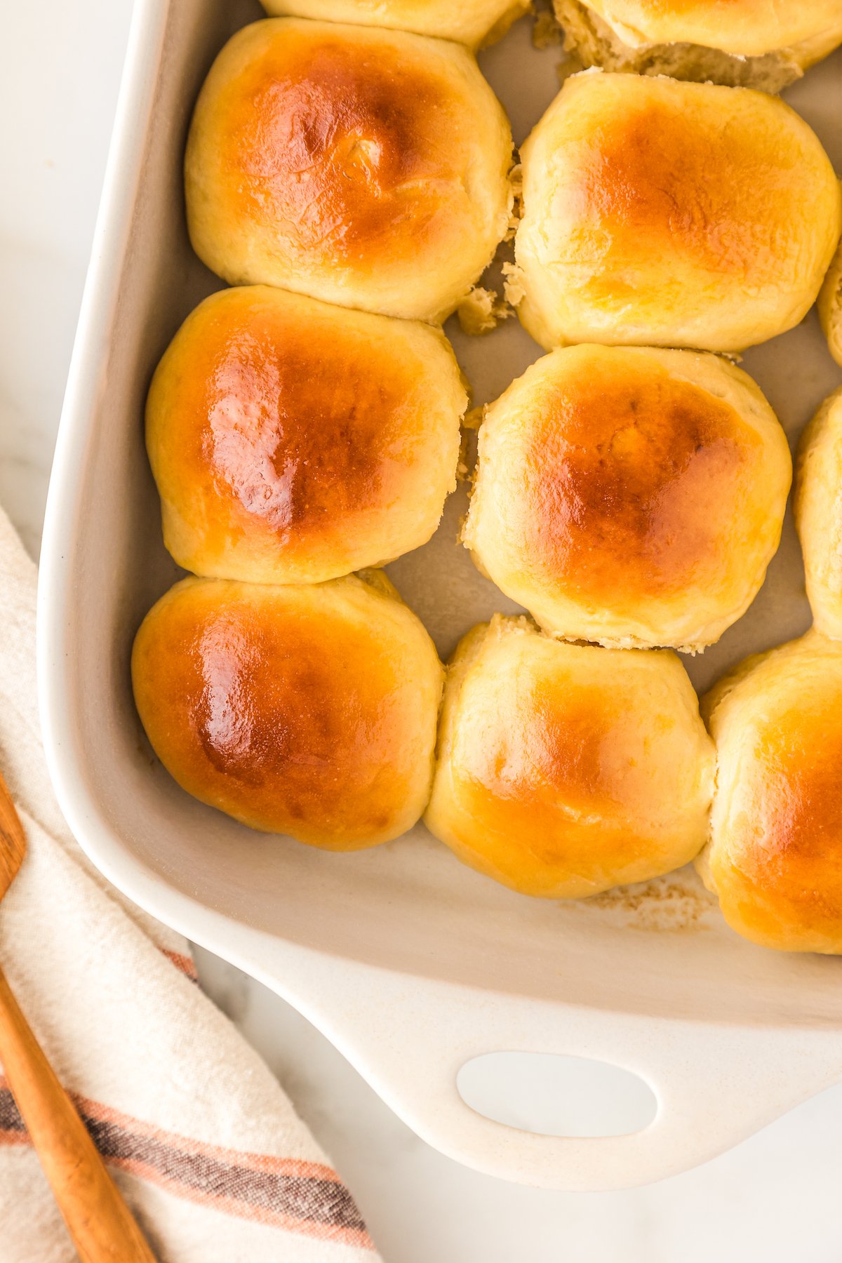 Soft & buttery Hawaiian Rolls are golden in color with a sweet hint of pineapple flavor. This homemade yeast roll recipe is a crowd favorite! Golden brown Hawaiian rolls are arranged closely together in a white baking dish. A striped kitchen towel and a wooden spoon are partially visible next to the dish on a light surface.