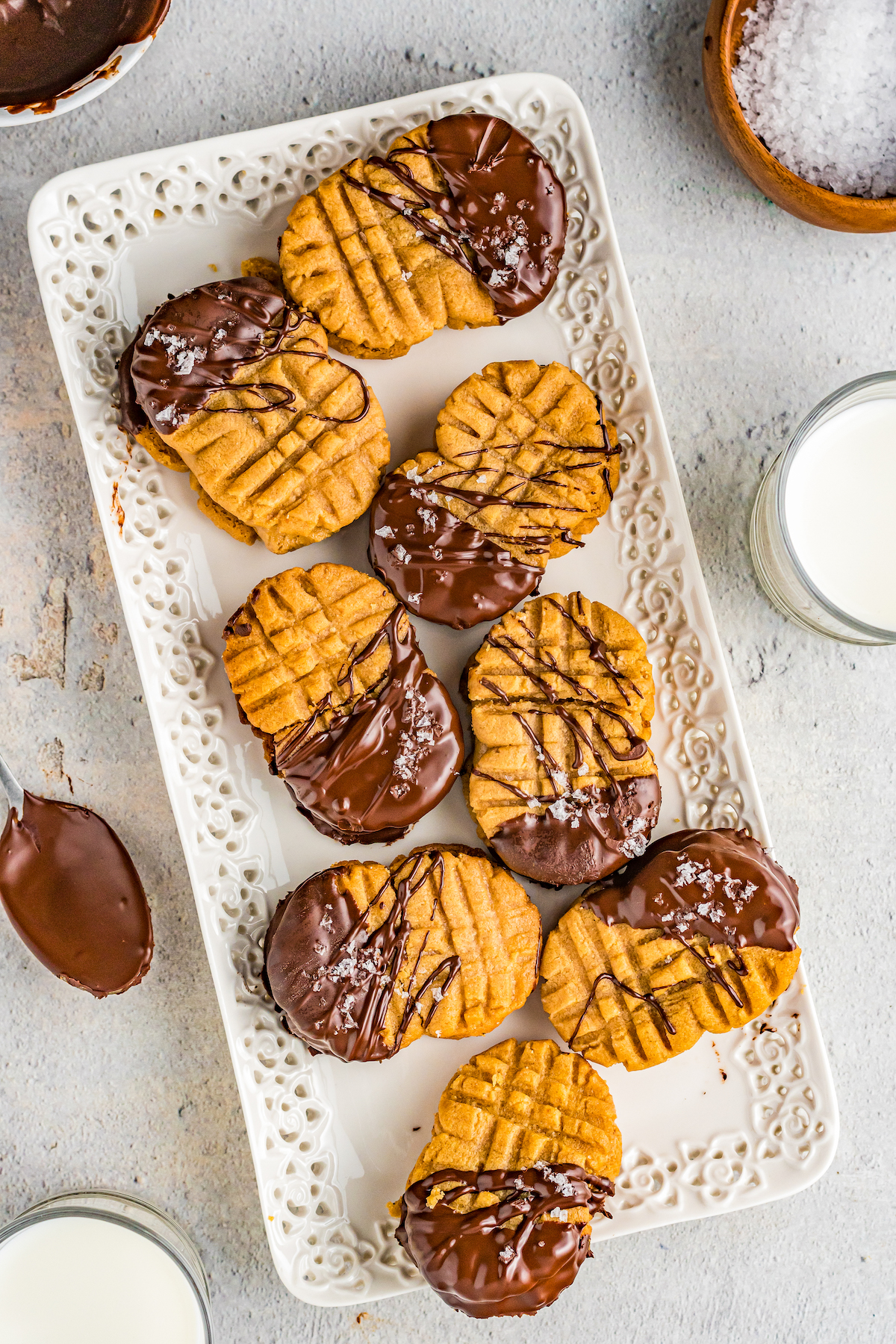 Chocolate-dipped homemade Nutter Butter cookies scattered on a white rectangular serving plate, with a glass of milk and chocolate-dipped spoon nearby.