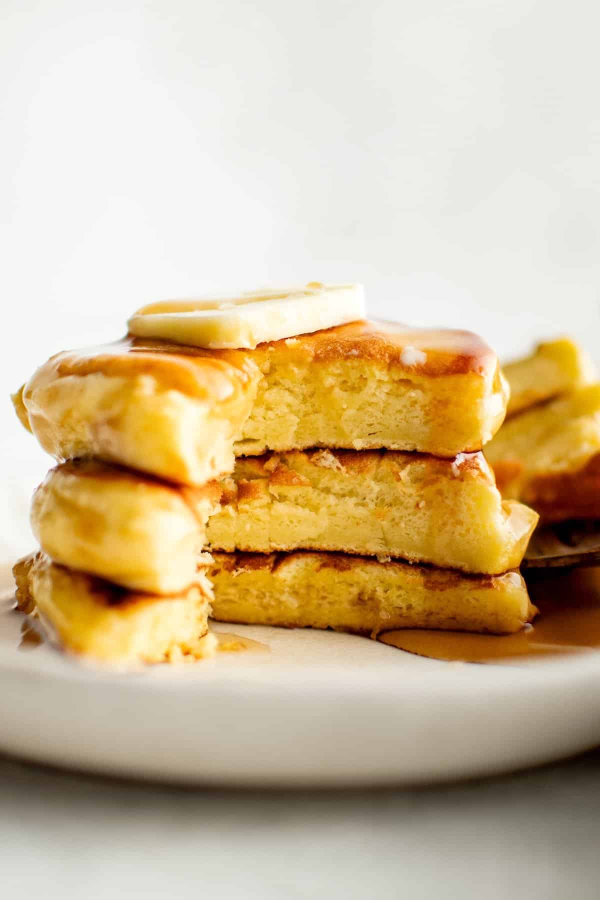 Ready in just 30 minutes, this light and fluffy Japanese Soufflé Pancake recipe is easy to make — with no special tools or molds required! A stack of three fluffy Japanese soufflé pancakes topped with a slice of butter and drizzled with syrup, on a white plate. A bite is taken out of the pancakes, revealing their soft, light and airy interior.