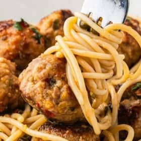 A close-up of chicken piccata meatballs and spaghetti twirled around a fork, garnished with herbs.