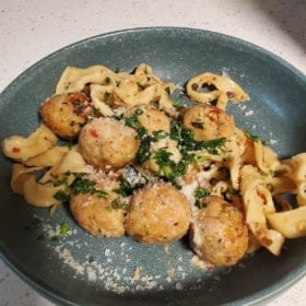 A plate containing several chicken meatballs on top of wide, twisted pasta noodles, garnished with chopped parsley and grated cheese.