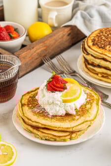 A serving of lemon ricotta pancakes, garnished with fresh fruit and whipped cream, on a breakfast table with syrup and other items.