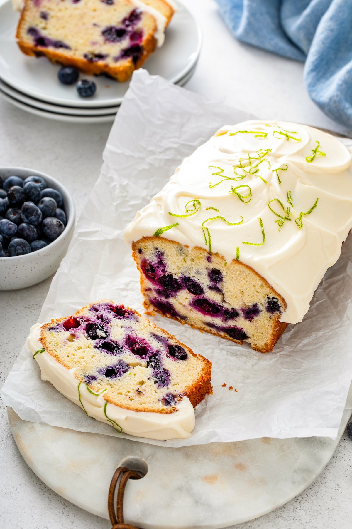 This moist and tender Lime Blueberry Pound Cake is bursting with fresh blueberries and lime zest with a thick cream cheese frosting on top! Lime Blueberry Pound Cake topped with creamy frosting and fresh lime zest being sliced into pieces on a parchment paper lined cutting board.