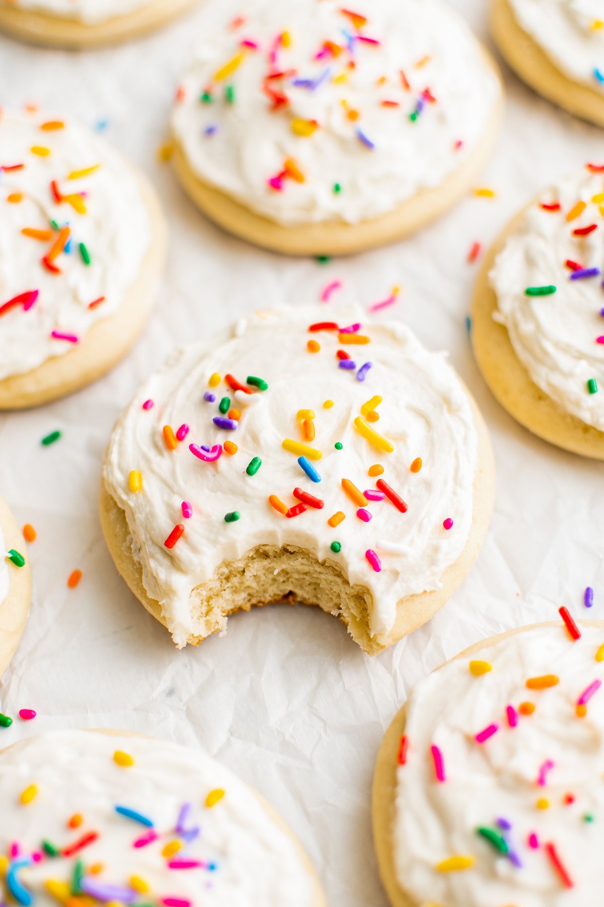 This copycat recipe for melt-in-your-mouth Lofthouse Cookies with homemade frosting are soft, thick and topped with fun sprinkles! Several Lofthouse cookies on a baking sheet. One has a bite taken out of it.