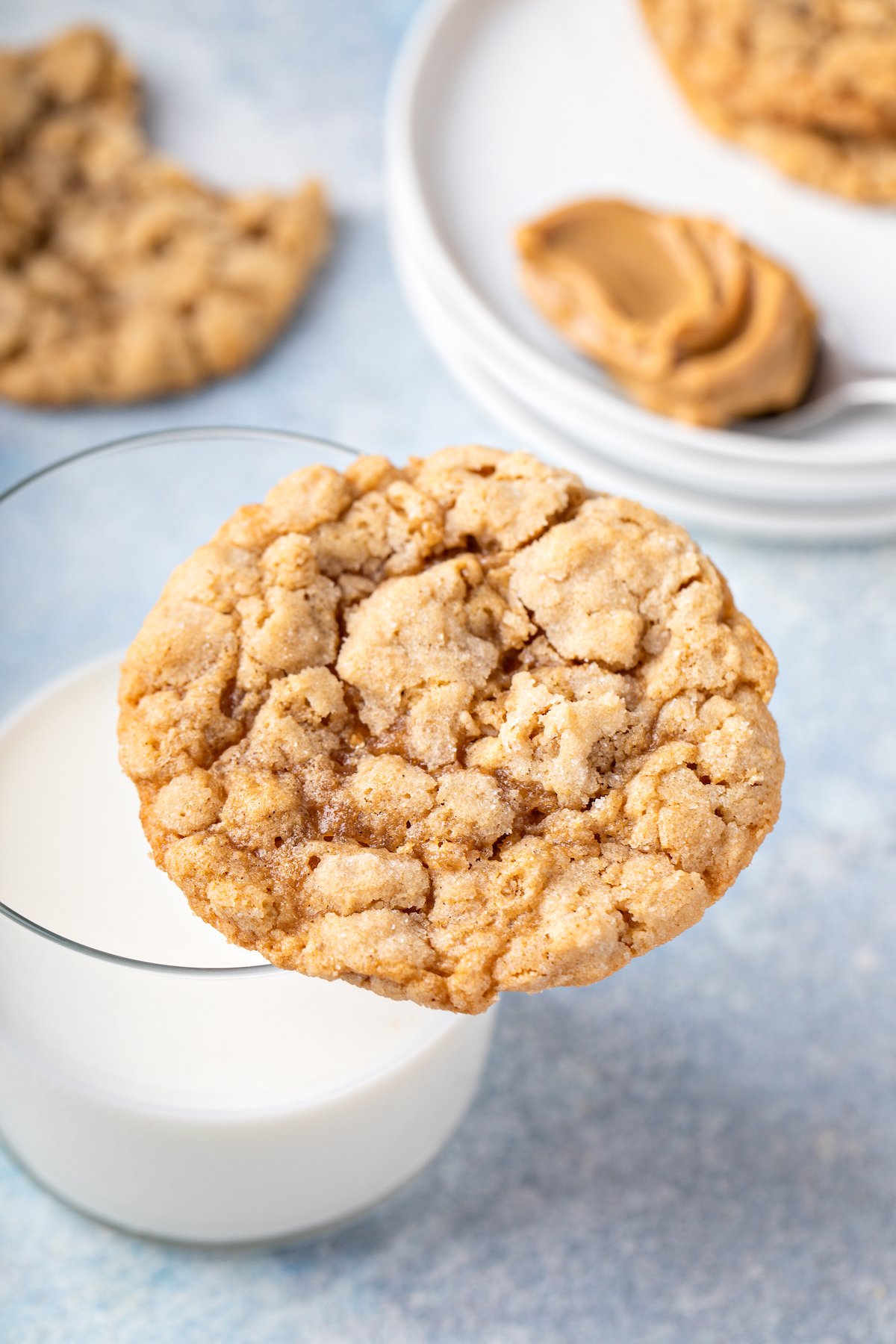 With a hint of cinnamon, crisp edges, and a chewy center, these wholesome Peanut Butter Oatmeal Cookies are sure to please! A peanut butter cookie balanced on the rim of a glass of milk. A saucer with pieces of cookie is in the background.