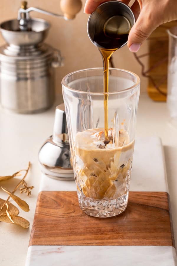 Smooth, spiced, and perfectly creamy, this Pumpkin Espresso Martini fall in a glass. Made with real pumpkin, espresso, and RumChata! A hand pours maple syrup from a small metal cup into a tall glass filled with ice, espresso, pumpkin, and milk on a wooden board, with a metal shaker and kitchen items in the background.