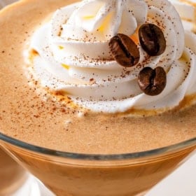 A close-up of a pumpkin espresso martini in a glass, topped with a swirl of whipped cream, coffee beans, and a sprinkle of spices.