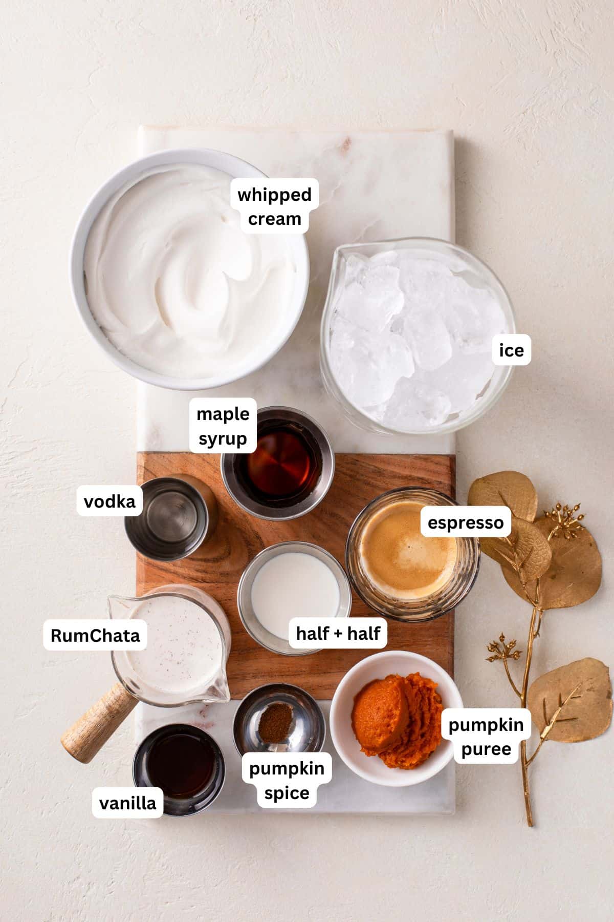 Smooth, spiced, and perfectly creamy, this Pumpkin Espresso Martini fall in a glass. Made with real pumpkin, espresso, and RumChata! A flat lay of ingredients labeled for a pumpkin martini recipe: whipped cream, ice, vodka, RumChata, maple syrup, half & half, espresso, pumpkin puree, pumpkin spice, and vanilla, arranged on a marble and wood board.