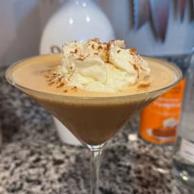 A creamy, brown pumpkin espresso martini in a martini glass topped with whipped cream and a sprinkle of cinnamon.