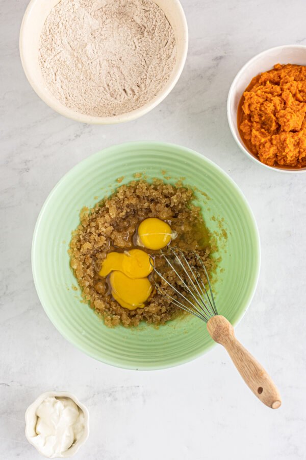 This bakery-style easy pumpkin muffin recipe creates moist and tender muffins made with a whole can of pumpkin and warm autumn spices! Brown sugar and eggs in a mixing bowl. A dish of pumpkin puree and a dish of dry ingredients are nearby on the work surface.