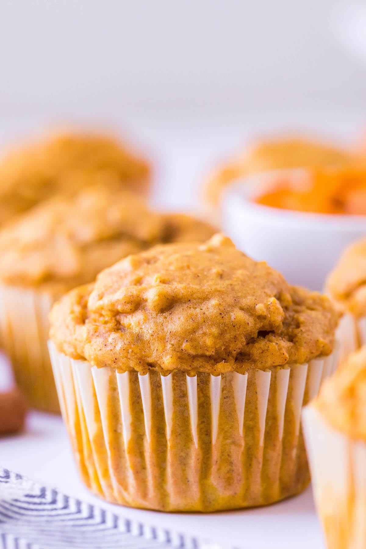 These Pumpkin Oatmeal Muffins are made easily in just one-bowl and are loaded with pumpkin flavor with a deliciously chewy consistency! Up close image of a pumpkin oat muffin surrounded by other muffins on a tea towel.