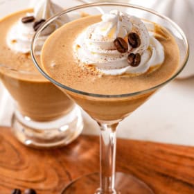 Two pumpkin spice espresso martinis in martini glasses topped with whipped cream, coffee beans, and a sprinkle of spice, set on a wooden surface with a blurred background.