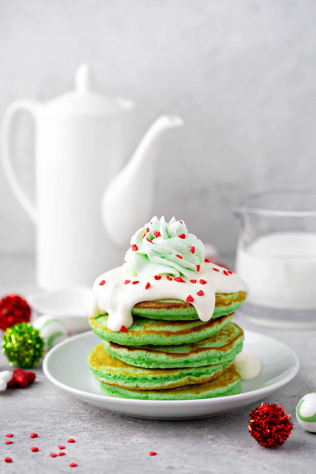 These fluffy Grinch Pancakes with cream cheese frosting are an easy homemade Christmas pancake recipe for a festive Christmas breakfast! A stick of red and green Christmas pancakes with red heart sprinkles and cream cheese frosting on top.