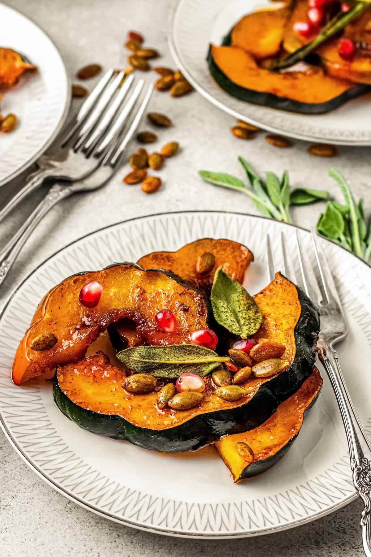 Sweet and spicy with perfectly caramelized edges, this Roasted Acorn Squash is the ultimate fall side dish topped with crispy pumpkin seeds. Slices of roasted acorn squash topped with pomegranate seeds, toasted pumpkin seeds, and fresh sage leaves are served on a white plate with a fork next to the dish.