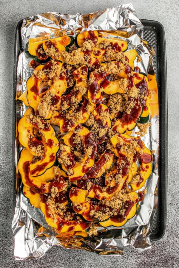Sweet and spicy with perfectly caramelized edges, this Roasted Acorn Squash is the ultimate fall side dish topped with crispy pumpkin seeds. Sliced acorn squash on a foil-lined baking sheet, topped with brown sugar and drizzled with a spiced sauce mixture, ready to be baked.