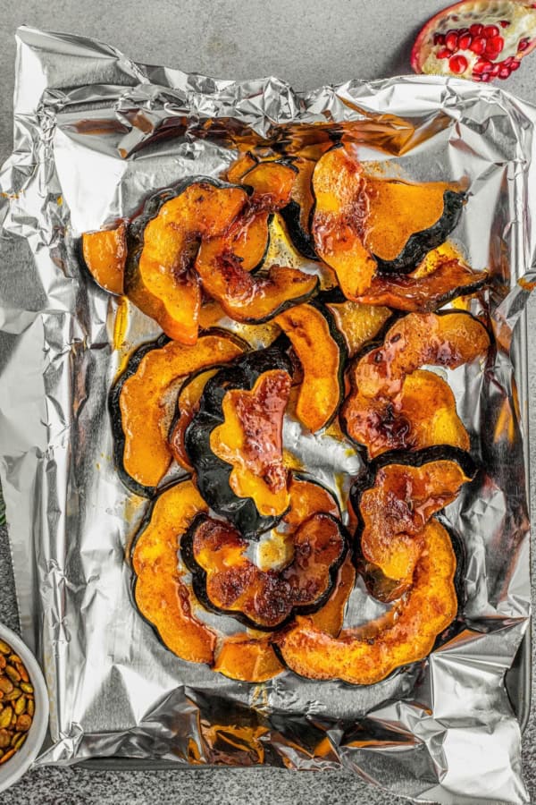 Sweet and spicy with perfectly caramelized edges, this Roasted Acorn Squash is the ultimate fall side dish topped with crispy pumpkin seeds. Sliced roasted acorn squash with caramelized edges on a foil-lined baking sheet, viewed from above. Some seeds and part of a pomegranate are visible in the corners.
