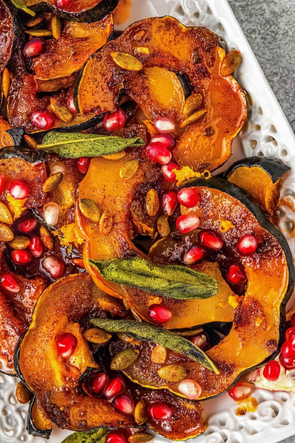 Sweet and spicy with perfectly caramelized edges, this Roasted Acorn Squash is the ultimate fall side dish topped with crispy pumpkin seeds. Sliced roasted acorn squash garnished with crispy sage leaves, pumpkin seeds, and bright pomegranate arils, served on a decorative white platter.