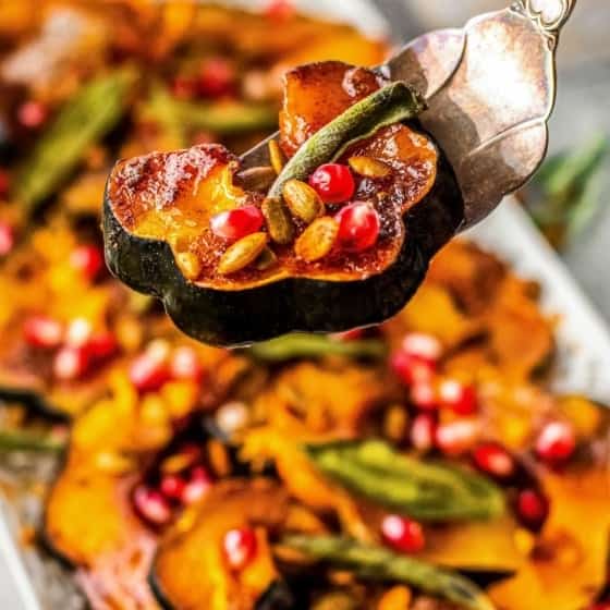 A close-up of roasted squash garnished with pomegranate seeds, pumpkin seeds, and fresh herbs on a silver serving utensil, with more squash on a platter in the background.