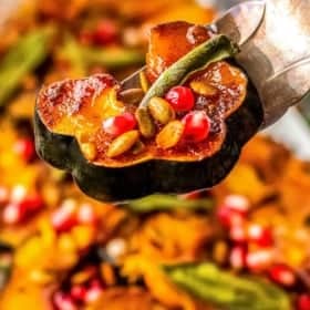 A close-up of tongs holding a slice of roasted acorn squash topped with pomegranate seeds and pumpkin seeds, with more squash pieces in the blurred background.