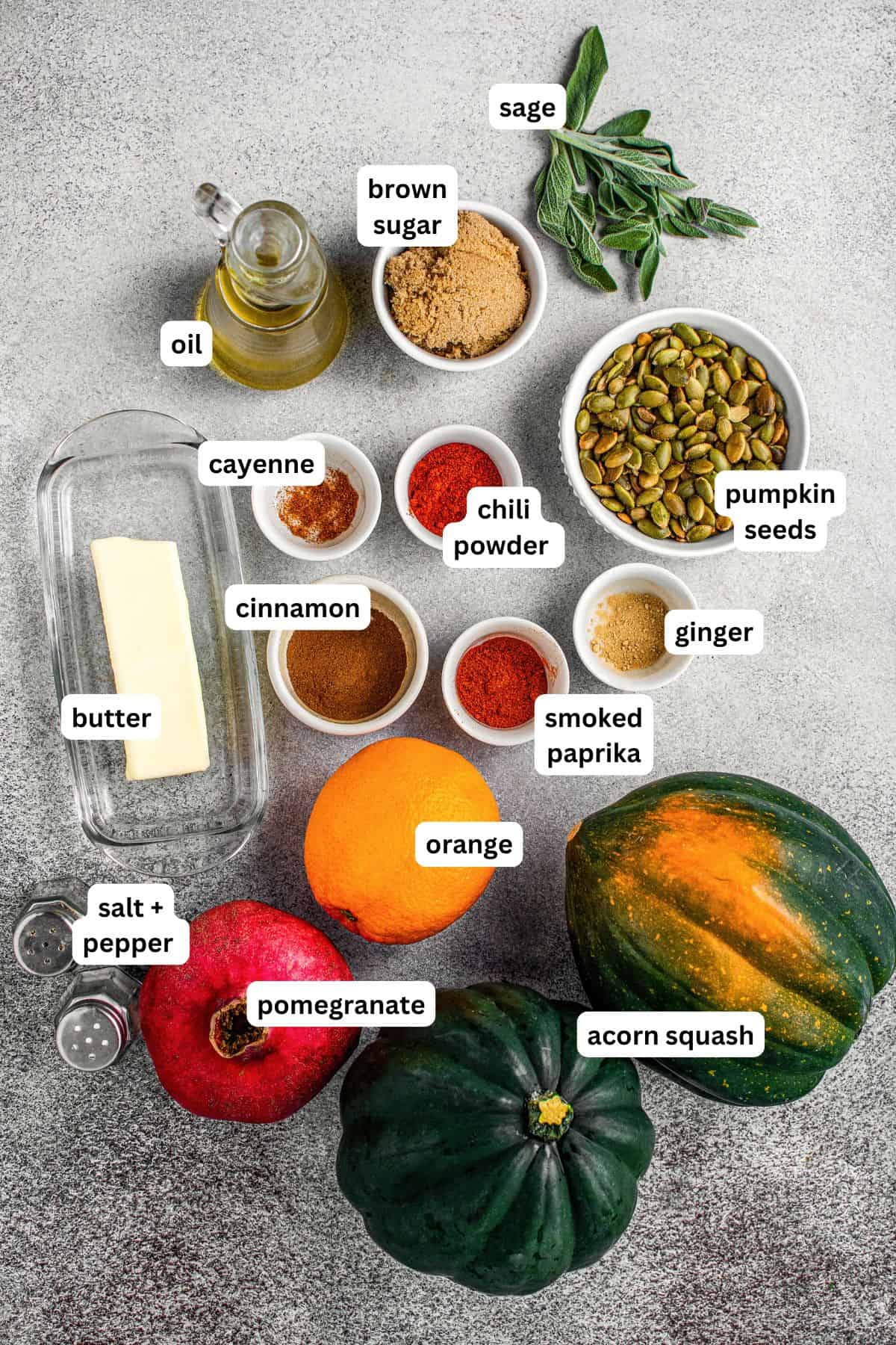 Sweet and spicy with perfectly caramelized edges, this Roasted Acorn Squash is the ultimate fall side dish topped with crispy pumpkin seeds. Overhead view of labeled ingredients for roasted acorn squash recipe on a gray surface: sage, oil, brown sugar, pumpkin seeds, ginger, smoked paprika, chili powder, cayenne, cinnamon, butter, salt, pepper, orange, pomegranate, and two acorn squash.