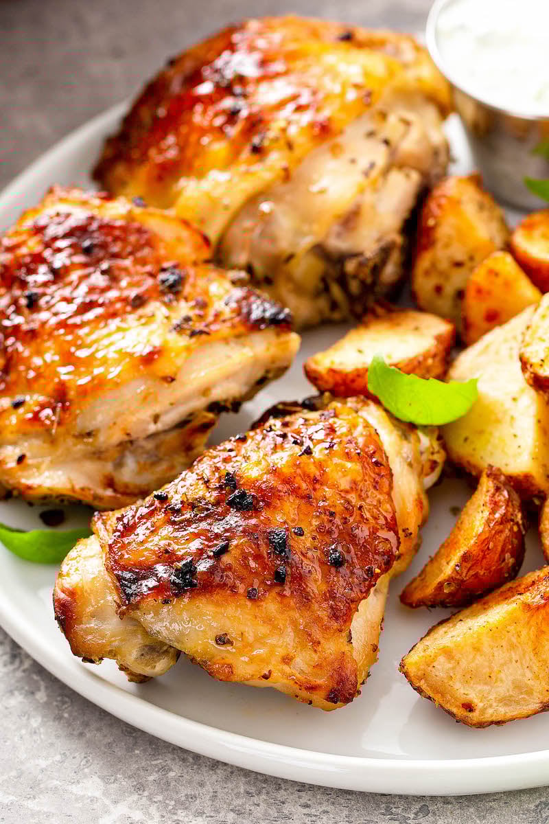 This is the best oven baked chicken recipe! Bone-in chicken breasts or thighs marinated and baked in the oven with a garlicky, citrus glaze. Baked chicken on a white plate with roasted potatoes.