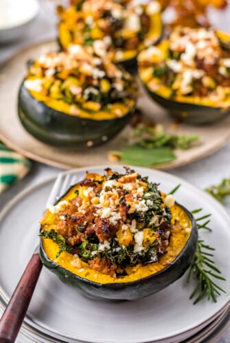A serving of cooked stuffed acorn squash, with a platter of stuffed acorn squash in the background. Fresh herbs decorate the plates and a fork lies nearby.