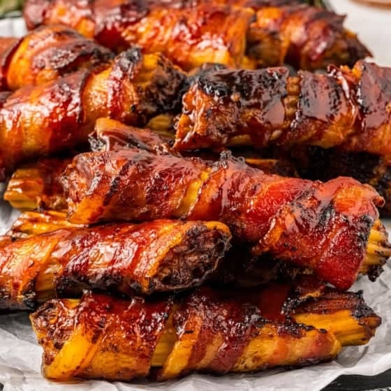 Close-up of a tray filled with bacon-wrapped stuffed shotgun shells, glazed with a shiny barbecue sauce. They are neatly arranged on crinkled parchment paper, with a checkered cloth napkin visible in the background.