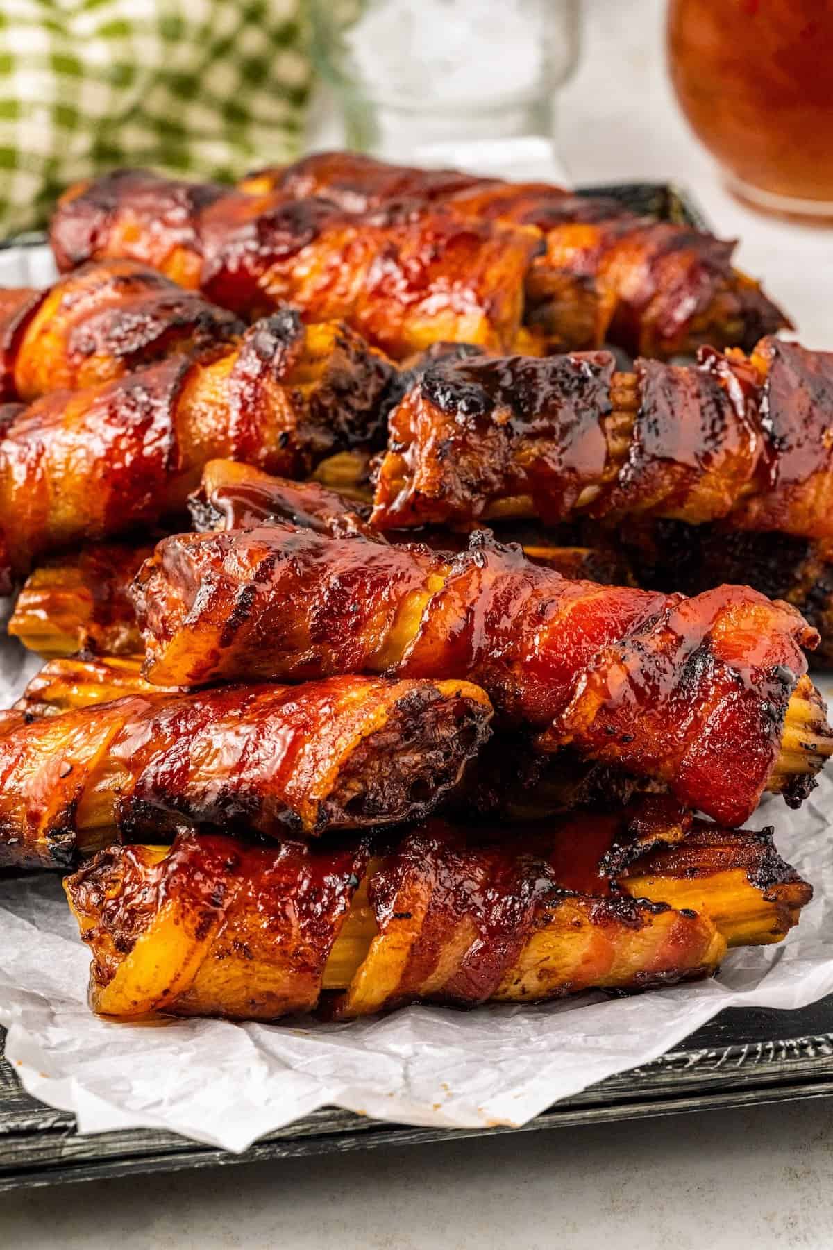 This mouthwatering bacon-wrapped BBQ Shotgun Shells recipe is the perfect smoky, cheesy, and meaty appetizer your guests will devour! Close-up of a tray filled with bacon-wrapped stuffed shotgun shells, glazed with a shiny barbecue sauce. They are neatly arranged on crinkled parchment paper, with a checkered cloth napkin visible in the background.