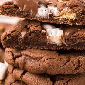 A stack of thick, chocolate smores cookies, filled with melted marshmallow and chocolate pieces, with one broken cookie on top revealing the gooey filling.