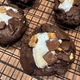 A close-up of chocolate s'mores cookies with toasted marshmallow, pieces of Hersheys chocolate, and graham crackers on top, cooling on a black wire rack over a wooden surface.