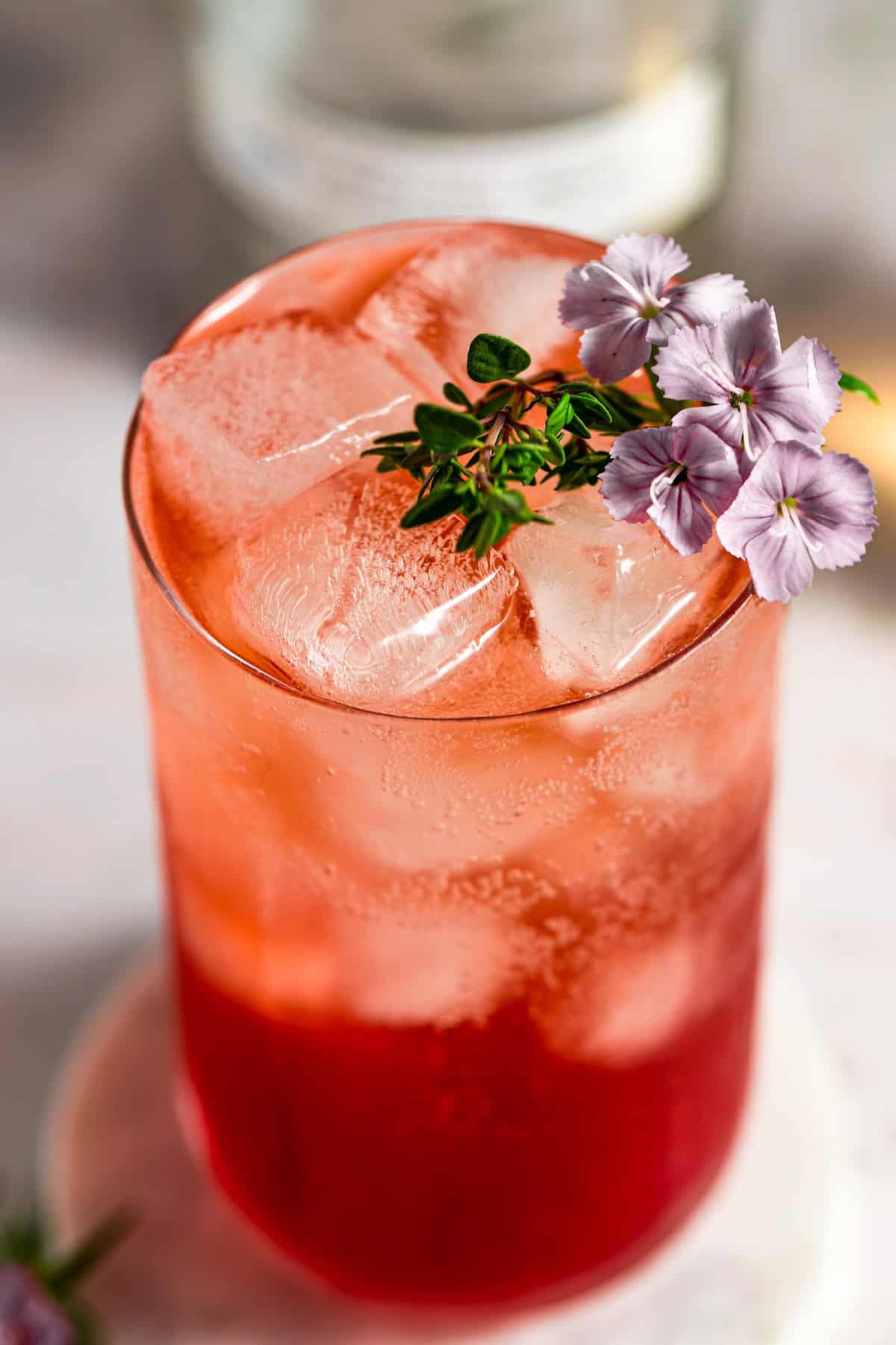 Fizzy, fruity, and refreshingly simple, this Transfusion Drink recipe is the best summer cocktail with vodka, grape juice, lime & ginger ale. A vibrant red iced transfusion drink in a glass, garnished with small purple flowers and a sprig of greenery, sits on a light surface. Large ice cubes fill the glass, and the background is softly blurred.
