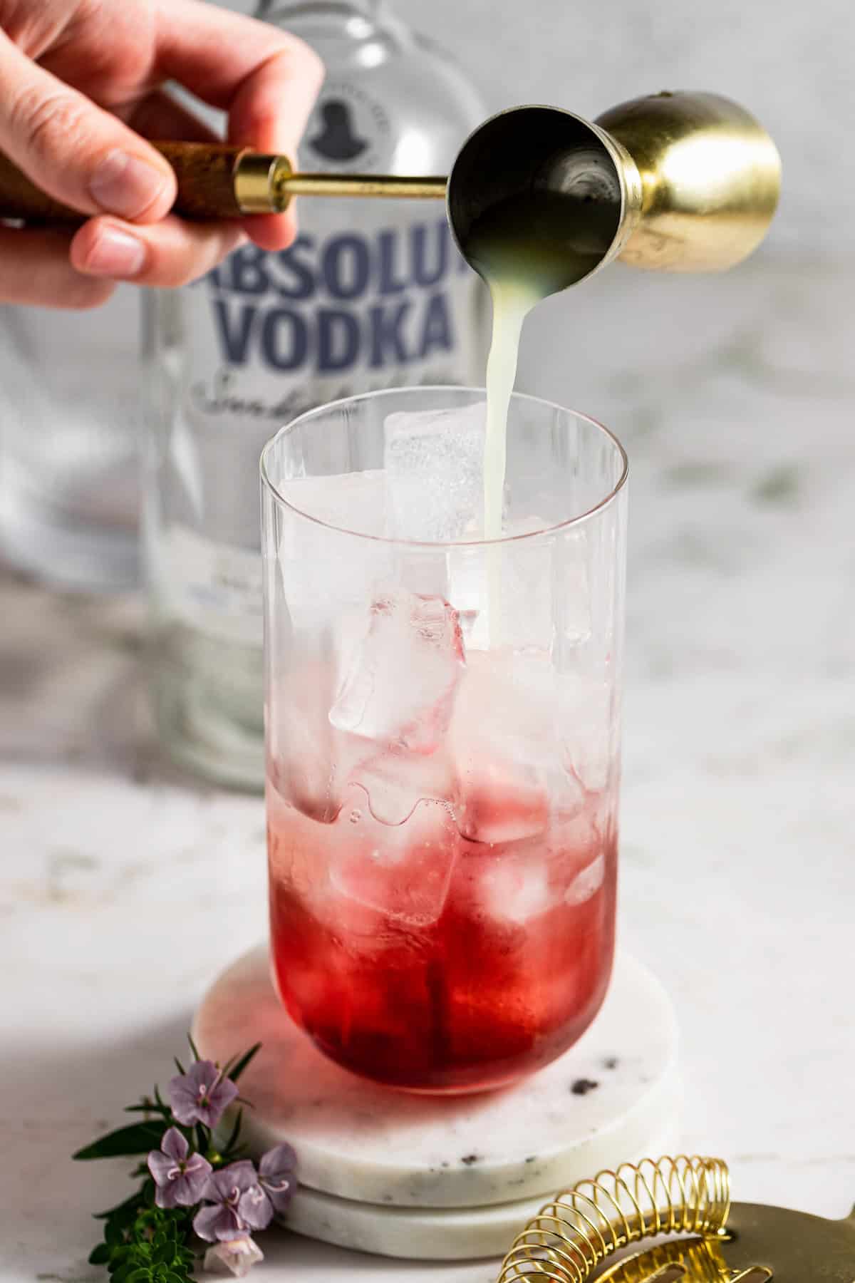 Fizzy, fruity, and refreshingly simple, this Transfusion Drink recipe is the best summer cocktail with vodka, grape juice, lime & ginger ale. A hand pours lime juice from a jigger into a glass filled with ice and grape juice, with a bottle of vodka in the background and small purple flowers and a gold strainer nearby.