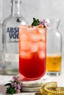 A tall glass of red and pink transfusion drink cocktail with ice, garnished with small purple flowers and herbs, sits on a marble coaster. A vodka bottle, a drink pitcher, and a cocktail strainer are in the background.