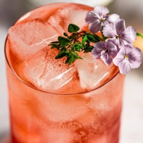 A glass of iced vodka transfusion drink, garnished with purple flowers and green herbs, displayed against a white background.
