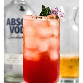 A vibrant red vodka transfusion cocktail with ice, garnished with purple flowers and herbs, sits in front of a vodka bottle and a glass with amber liquid.